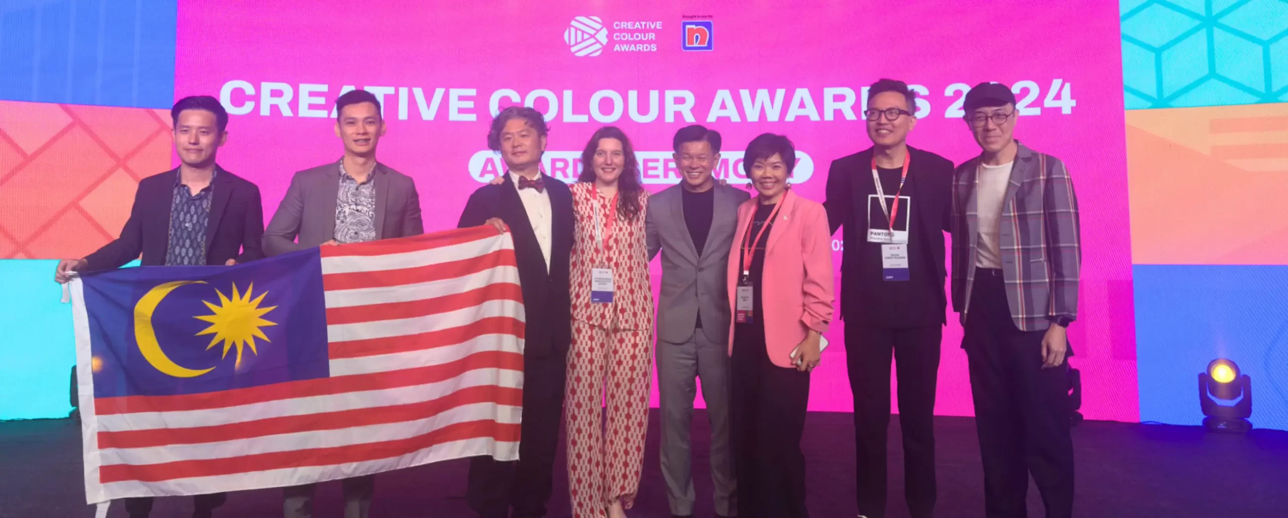 Group photo at the Creative Colour Awards 2024, showcasing diverse attendees including Malaysian representatives holding the national flag. The vibrant background highlights Nippon Paint branding, reflecting creativity and innovation in design.