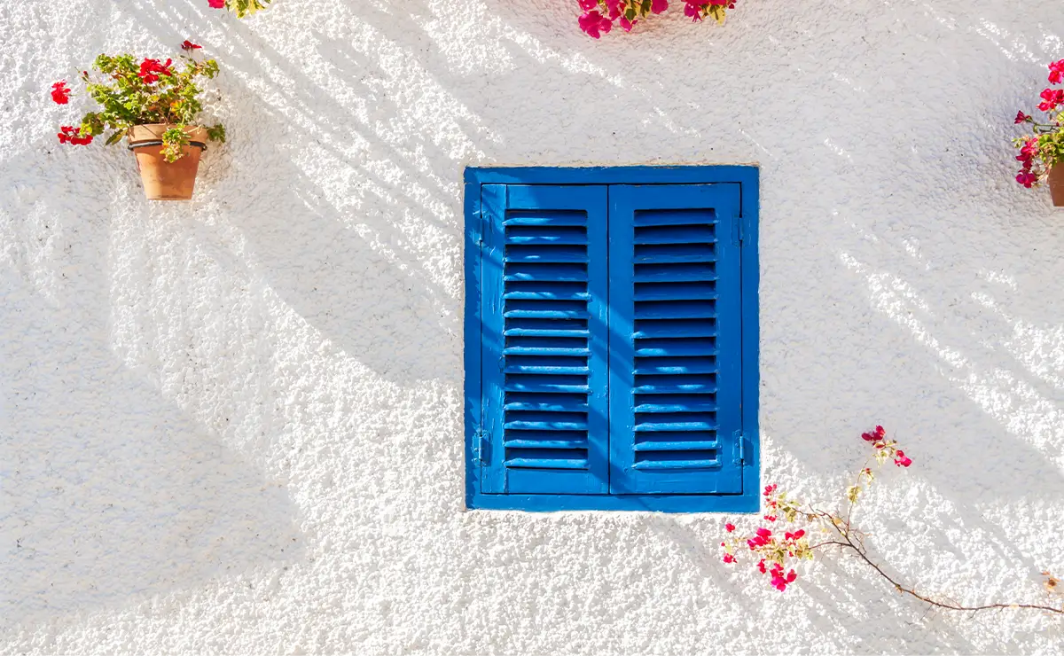 Bright blue wooden shutters on a white wall adorned with vibrant flowers, showcasing a charming Mediterranean aesthetic. The use of Nippon Paint products ensures a durable and weather-resistant finish, enhancing the visual appeal of this outdoor space.