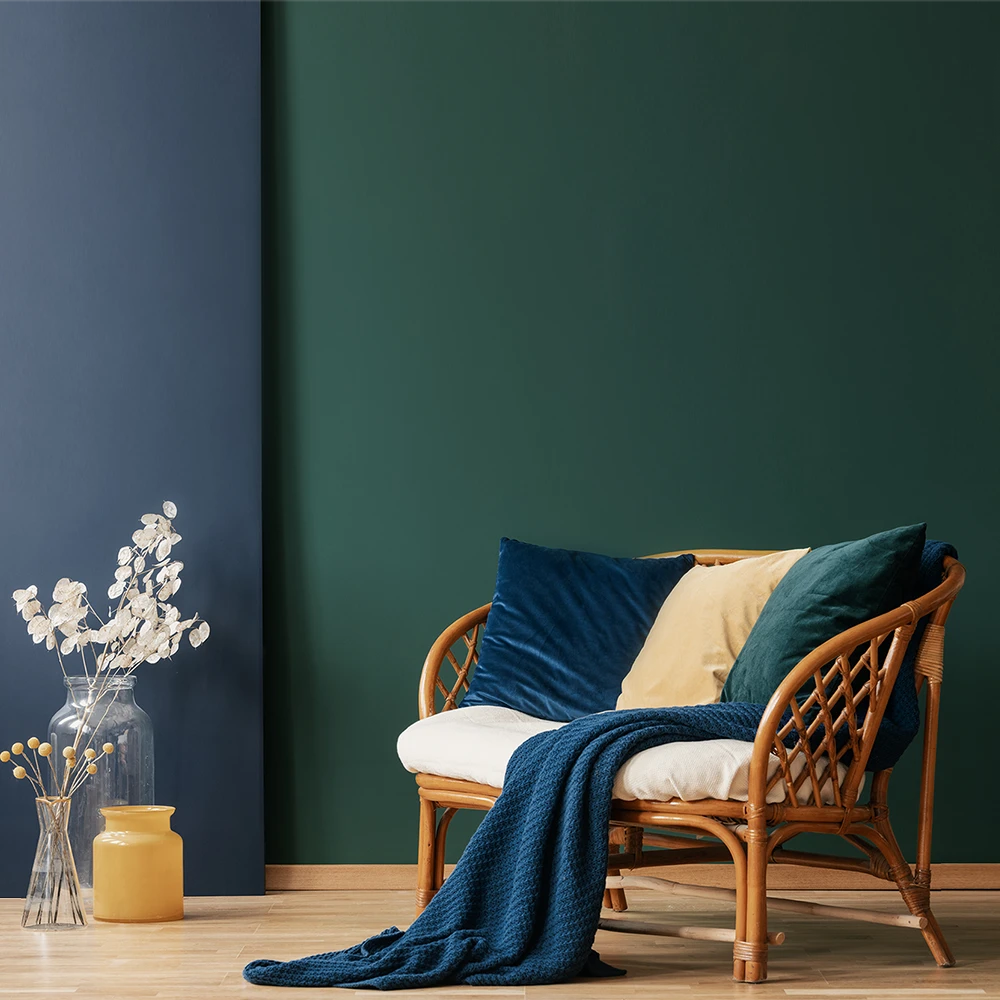 Cozy living space featuring a rattan sofa adorned with colorful pillows, surrounded by rich green and deep blue walls painted with Nippon Paint products, complemented by decorative vases and a warm throw blanket.